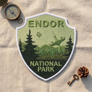 May include: A shield-shaped embroidered patch featuring a light green background with dark green pine trees, mountains, a satellite dish, and a small circular object. The text reads ENDOR NATIONAL PARK in dark green block letters.