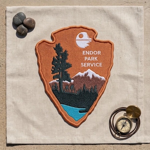 May include: An embroidered patch shaped like a shield with an orange background, featuring a dark green tree, brown mountains with white peaks, a blue lake, and a white Death Star icon. The text reads ENDOR PARK SERVICE in white letters.