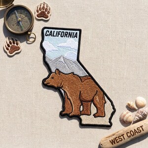 May include: An embroidered patch shaped like the state of California featuring a brown bear, mountain range, and clouds with the text CALIFORNIA. Includes a brass compass, two bear paw patches, three seashells, and a piece of wood labeled WEST COAST.