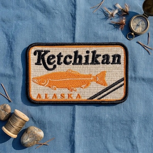May include: A rectangular embroidered patch featuring the word Ketchikan in black text above an orange fish illustration, with the word Alaska in orange text below, all set against a beige background with a black and orange border trim.