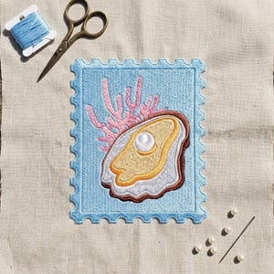 May include: An embroidered patch shaped like a postage stamp featuring a light blue background, a brown and yellow oyster shell with a white pearl, and pink coral. Includes a pair of metal scissors, a spool of light blue thread, and a needle.
