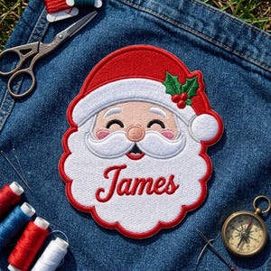 May include: An embroidered patch featuring a smiling Santa Claus face with a red hat, white beard, and green holly leaves. The name James is stitched in red cursive script across the white beard. The patch is placed on blue denim fabric.