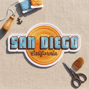May include: A circular embroidered patch featuring a yellow and orange sunburst design with the text SAN DIEGO in light blue block letters and California in brown script. The patch is surrounded by sewing tools including thread, a needle, and scissors.