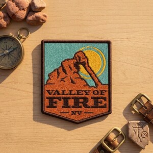 May include: A rectangular embroidered patch featuring a stylized orange rock formation against a light blue sky with a yellow sun. The text VALLEY OF FIRE NV is stitched in dark brown across the lower portion of the patch, which has a dark brown border.
