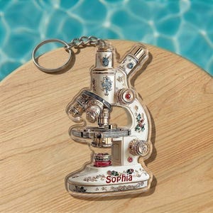 May include: A white and gold microscope keychain with the name "Sophia" printed on the base. The keychain has a silver chain and ring. The microscope has decorative floral and geometric designs.