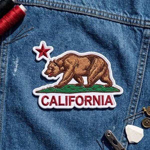 May include: An embroidered patch featuring a brown grizzly bear walking on a green patch of grass, a red star above, and the word CALIFORNIA in red capital letters, all outlined in white, placed on blue denim fabric.