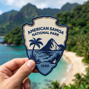 May include: An embroidered patch shaped like a shield with a cream background and dark blue border. It features a palm tree, a mountain, and the text AMERICAN SAMOA NATIONAL PARK above the year 1988 in dark blue thread against a blue landscape.