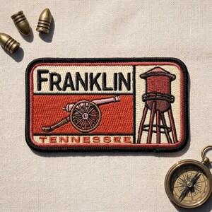 May include: A rectangular embroidered patch featuring the text FRANKLIN TENNESSEE in black, a brown cannon, and a brown water tower on a cream and rust-colored background. Three brass bullet casings are positioned above, and a brass compass below.