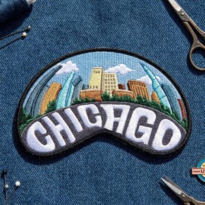 May include: A semi-circular embroidered patch featuring a stylized Chicago skyline with tan, brown, and blue buildings against a light blue sky with white clouds. The word CHICAGO is stitched in large white letters across a dark gray curved base.