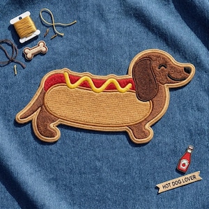 May include: An embroidered patch of a brown dachshund shaped like a hot dog with red and yellow thread details. Includes a small bone patch, a spool of gold thread, a ketchup bottle patch, and a banner patch with the text HOT DOG LOVER.