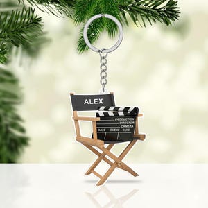 May include: A keychain featuring a miniature director's chair and clapperboard. The chair has the name "ALEX" on the back. The clapperboard includes the words "PRODUCTION, DIRECTOR, CAMERA, DATE, SCENE, TAKE". The keychain is attached to a silver keyring.