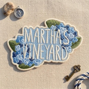 May include: An embroidered patch featuring the text MARTHA'S VINEYARD in white thread against a light blue background, surrounded by clusters of blue hydrangea flowers and green leaves, with a cream-colored border and a scalloped edge.