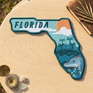 May include: An embroidered patch shaped like the state of Florida featuring the text FLORIDA in dark teal. The design includes a light blue sky, a peach-colored sun, white clouds, dark teal palm trees, and a light blue dolphin in the ocean.