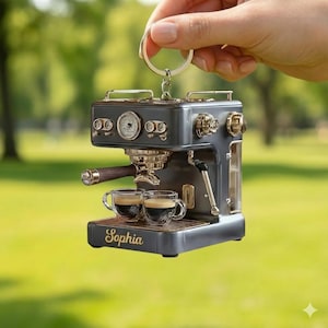 May include: Miniature espresso machine keychain in gray and gold tones, with two small glass cups. The word "Sophia" is inscribed on the base. The keychain is held up by a hand against a blurred green background.