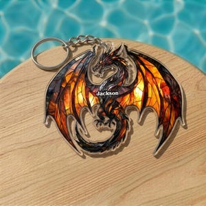 May include: A dragon-shaped keychain with a stained-glass effect. The dragon has orange, yellow, and black wings, a red head, and the name "Jackson" printed on its body. The keychain is attached to a silver chain and ring.