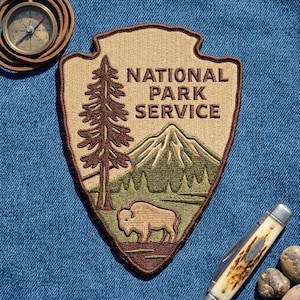 May include: An embroidered National Park Service patch in the shape of an arrowhead, featuring a brown pine tree, a green mountain, and a tan bison. The patch is displayed alongside a round brass compass and a folding pocket knife.