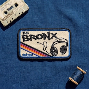 May include: A rectangular embroidered patch featuring the text THE BRONX and NEW YORK in black, a black line drawing of headphones, and a diagonal stripe pattern in red, orange, yellow, and blue on a cream background with a black border.