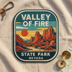 May include: A rectangular embroidered patch with rounded corners featuring a landscape of the Valley of Fire State Park in Nevada. The design includes red rock formations, a yellow sun, and blue sky, with the text VALLEY OF FIRE STATE PARK NEVADA.