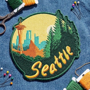 May include: A circular embroidered patch featuring the Seattle skyline with the Space Needle in orange, blue city buildings, and green pine trees against a yellow sun. The word Seattle is stitched in yellow cursive script across the bottom.