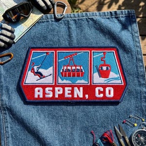 May include: A rectangular embroidered patch featuring three panels depicting a skier, a cable car, and a gondola against a light blue sky and white snow. The patch has a red border and a red base with the white text ASPEN, CO.