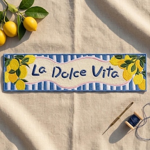 May include: A rectangular embroidered patch featuring a blue and white striped background with yellow lemon clusters on the left and right sides. The center displays the text La Dolce Vita in dark blue script, outlined by a light pink wavy border.