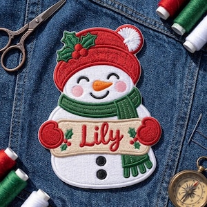 May include: An embroidered snowman patch featuring a red hat with a white pom-pom and green holly, a green scarf, and red mittens. The snowman holds a beige banner with the name Lily in red script, and has two black buttons on its body.