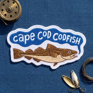 May include: An embroidered patch featuring a brown and tan codfish centered below the blue text "cape COD CODFISH" on a white background. The patch has a white border and is shaped like a speech bubble, showcasing detailed stitching of the fish.