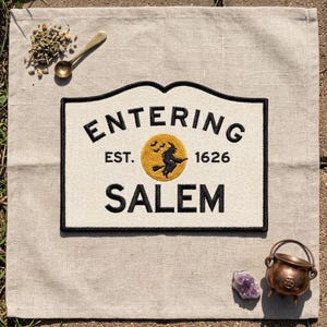 May include: A rectangular embroidered patch with a cream-colored background and a black border. The text reads ENTERING SALEM with EST. 1626 on either side of a central gold circle featuring a black silhouette of a witch flying on a broomstick.