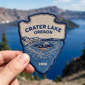 May include: An embroidered patch in the shape of a National Park Service arrowhead featuring a tan background with dark blue borders and text. The patch displays the words CRATER LAKE OREGON and 1902, with a blue lake and mountain landscape.