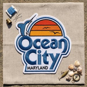May include: An embroidered patch featuring a blue marlin jumping over a sunset graphic with the text Ocean City MARYLAND in blue and black thread. The patch is surrounded by blue thread, a metal thimble, needles, and various small seashells.