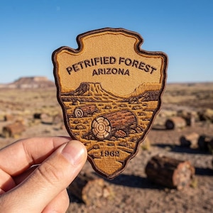 May include: A shield-shaped embroidered patch featuring a tan background with dark brown stitching. The text PETRIFIED FOREST ARIZONA is arched at the top, with a landscape of desert hills and fallen logs below, and the year 1962 at the bottom.