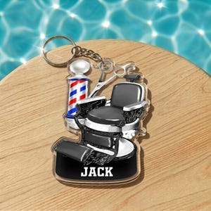 May include: A barber-themed keychain featuring a miniature barber chair, scissors, and a barber pole with red, white, and blue stripes. The keychain has the name "JACK" printed on it. The background is a wooden surface with a water reflection.