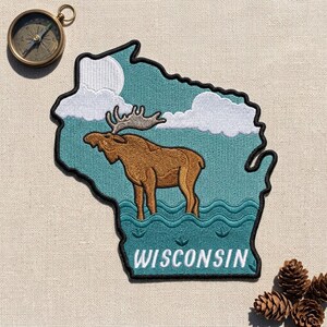 May include: An embroidered patch shaped like the state of Wisconsin featuring a brown moose standing in teal water with white clouds and a moon in the background. The word WISCONSIN is stitched in white at the bottom of the patch.