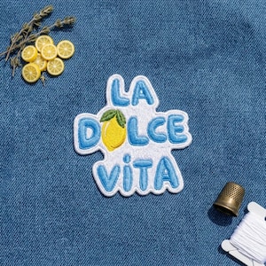 May include: An embroidered patch featuring the text LA DOLCE VITA in blue thread with a yellow lemon icon replacing the letter O. The patch has a white border and is placed on blue denim fabric alongside small lemon slices and a metal thimble.