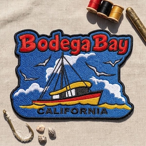 May include: An embroidered patch featuring a blue background with white clouds and seagulls. A fishing boat with a yellow hull and red trim is centered. The text Bodega Bay is in red at the top, and CALIFORNIA is in black at the bottom.