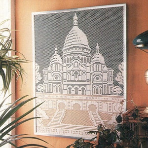 May include: A white crocheted wall hanging depicting a church with a dome and two towers. The hanging is framed in a silver frame and is displayed on a wall with a light orange color.