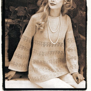 May include: A woman wearing a beige knitted sweater with a delicate lace pattern and a pearl necklace. The sweater has a relaxed fit with bell sleeves. The woman is sitting in front of a leafy background.