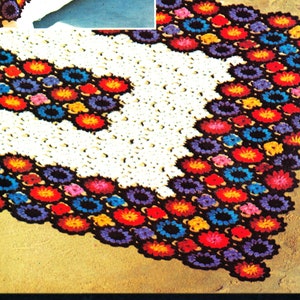 May include: A white crocheted throw blanket with a colorful floral pattern. The blanket is made with a variety of colors, including red, orange, yellow, green, blue, purple, and black. The blanket is shown on a bed and on the floor.