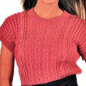 以下が含まれることがあります： 短い袖のピンクのケーブルニットセーターを着た女性。彼女は笑顔で、長いブロンドの髪をしています。