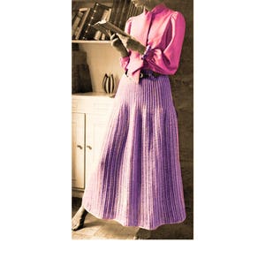 May include: A woman wearing a pink blouse with a high collar and a long, purple crochet skirt. She is standing in a room with a bookshelf and a vase on a table.
