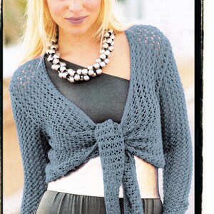 May include: A light blue crocheted shrug with long sleeves and a tie at the front. The shrug is worn over a black tank top and a dark gray skirt.