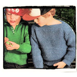 May include: Two children wearing knitted sweaters and hats. The child on the left is wearing a green sweater with a button closure and a red, white, and blue baseball cap. The child on the right is wearing a blue sweater and a black and white baseball cap.