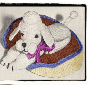 May include: A white knitted poodle dog toy with a purple bow, lying in a brown, blue, and yellow knitted dog bed.