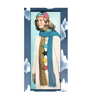 May include: A woman wearing a white coat and a blue and gold crocheted scarf with star-shaped embellishments. She is also wearing a multi-colored crocheted hat.