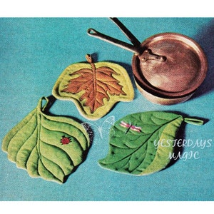 May include: Four quilted pot holders shaped like leaves, with embroidered details. The pot holders are green, brown, and yellow. The pot holders are on a blue background. The text "YESTERDAYS MAGIC" is on the bottom right corner of the image.