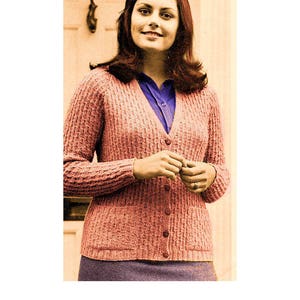Könnte beinhalten: Eine Frau trägt einen rosa gestrickten Cardigan mit Knöpfen. Der Cardigan hat ein strukturiertes Muster und wird über einem blauen Hemd getragen.