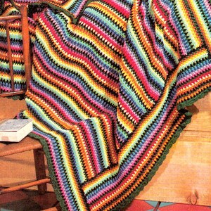 May include: A colorful, striped crochet blanket with a green border is draped over a bed and a wooden chair. The blanket features a vibrant array of colors, including red, yellow, blue, and green. A matching pillow and quilt are on the bed. A white hat rests on the chair.