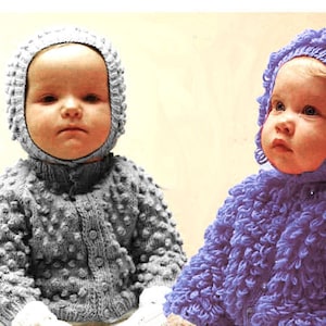 May include: Two babies wearing handmade knitted outfits. One baby is in a gray textured sweater with a matching hood and mittens. The other baby is in a purple textured sweater with a matching hood and mittens. Both babies are wearing white knitted pants.