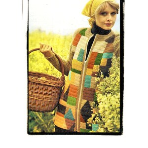 May include: A woman wearing a yellow headscarf and a colorful crocheted jacket stands in a field of yellow flowers. She is holding a wicker basket.