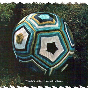 Peut inclure: Une balle en crochet avec un motif géométrique dans des tons de bleu, vert, blanc et noir. La balle est posée sur un lit d'herbe verte.